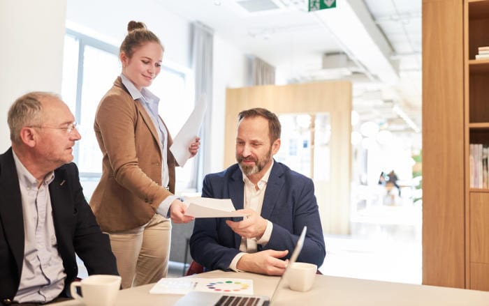 Junge Beraterin, die zwei Männern Unterlagen aushändigt: Die Gehälter von Auszubildenden im Versicherungsbereich liegen im ersten Jahr bei 1.205 Euro.