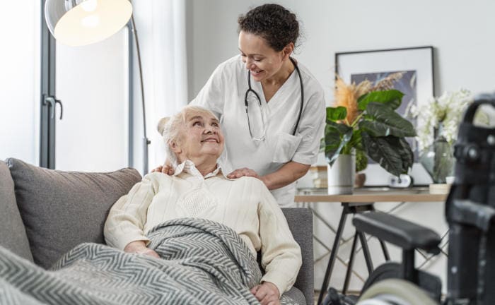 Ärztin redet mit älterer Patientin, die auf einem Sofa liegt: Eine Pflegeversicherung springt im Alter ein, wenn sich Versicherte nicht mehr selbst versorgen können.