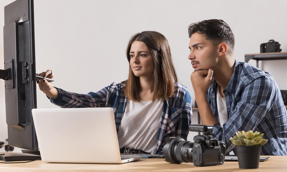 Frau erklärt Kollegen etwas am PC: Ihre Webseiten auf SEO zu optimieren, ist für Versicherer wichtig, damit Kunden auf Produkte und Dienstleistungen aufmerksam werden.